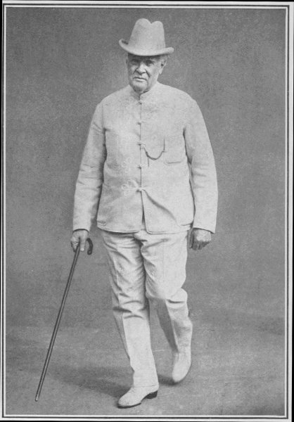 Zwart-wit foto van een witte man in een licht pak. Hij draagt een bijbehorende lichte hoed en lichte schoenen, en heeft een wandelstok vast.