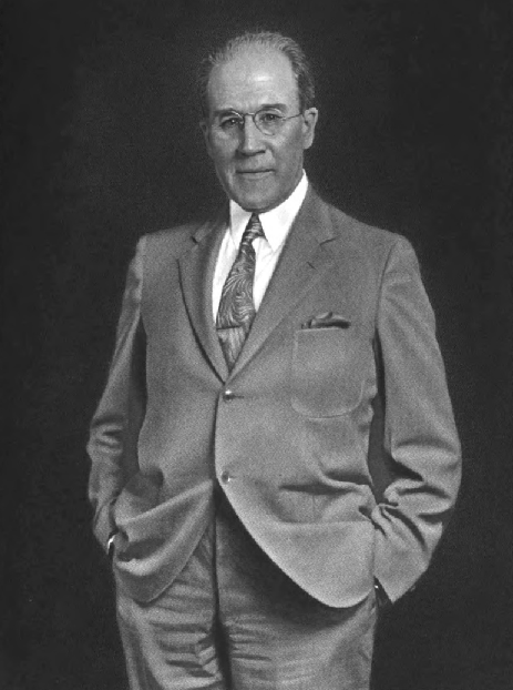 Zwart-wit foto van een witte, kalende man in een pak met een das. Hij draagt een bril, houdt beide handen in zijn broekzakken en kijkt recht in de camera.