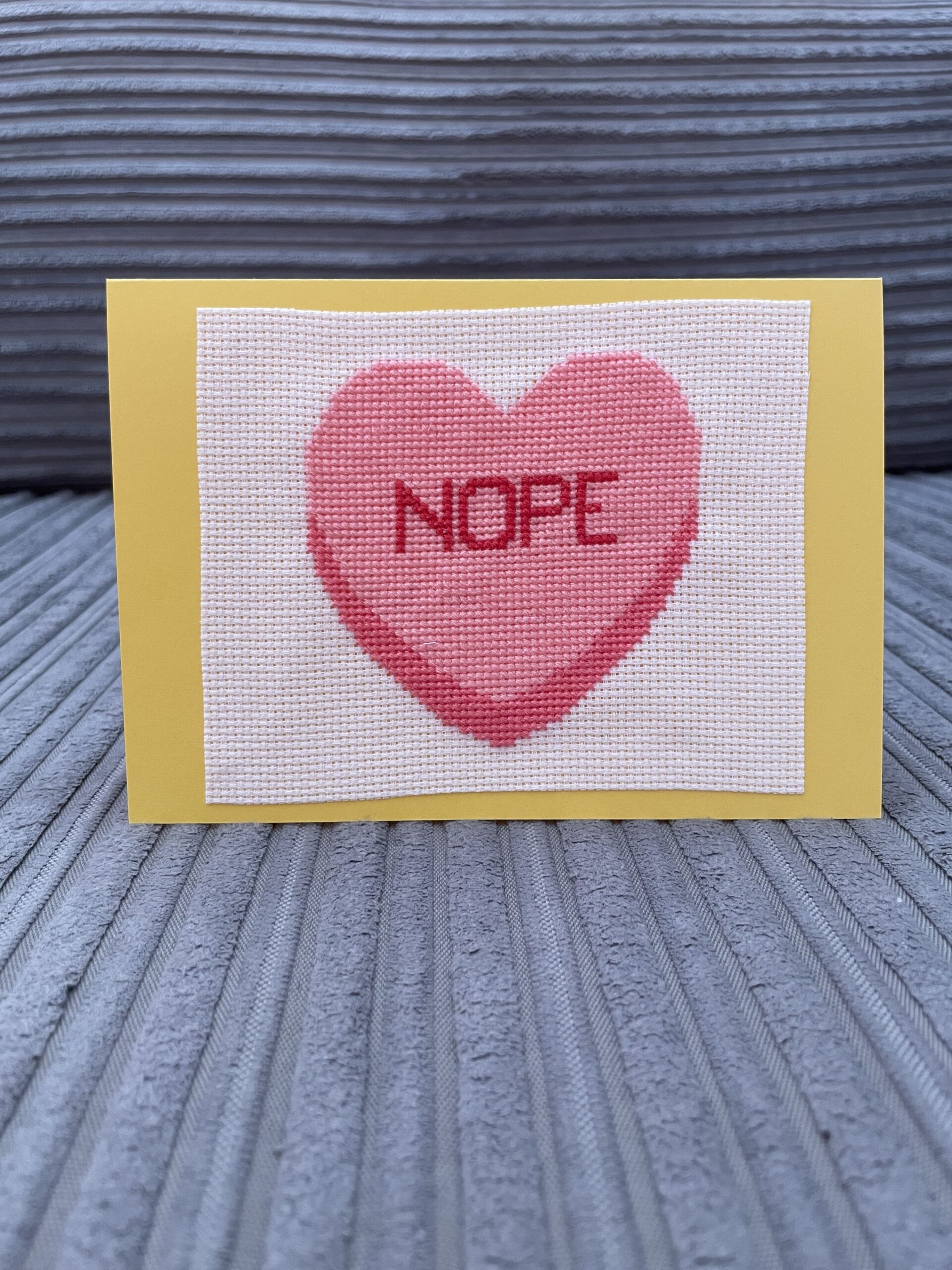 Een gele postkaart met daarop een cross stitch van een roze hart met 'nope' erin geschreven.