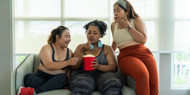 Drie dikke personen zitten naast elkaar op de sofa. De middelste houdt een doos popcorn vast, waar ze allemaal uit eten.